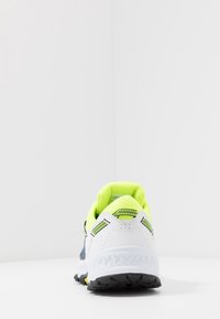 Laufschuh mit einem weißen Äußeren, fluoreszierenden gelben Akzenten, einem strukturierten Fersenkragen und einer schwarzen Gummisohle mit multidirektionalen Profilen.
