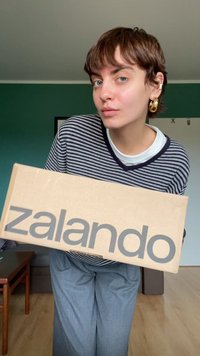 Pappkarton, gehalten von einer Person mit den fett gedruckten, grauen Buchstaben "zalando". Die Person trägt ein gestreiftes Oberteil und graue Hosen, vor einem neutralen Hintergrund.
