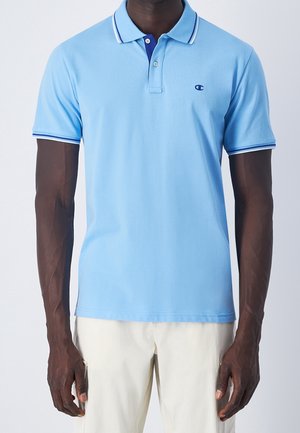 Mann trägt hellblaues kurzärmeliges Poloshirt mit gestreiftem Kragen und Manschetten, kombiniert mit hellen Hosen, steht vor einfarbigem Hintergrund.