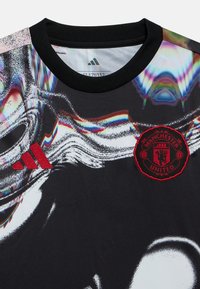 Camiseta gráfica negra con diseño texturizado, logo rojo de Adidas y escudo del Manchester United. Cuello redondo negro y mangas cortas.