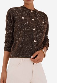 Cardigan lavorato a maglia marrone scuro con accenti bianchi maculati, colletto rotondo, chiusura frontale con bottoni, polsini e orlo a costine. Indossato sopra pantaloni chiari.