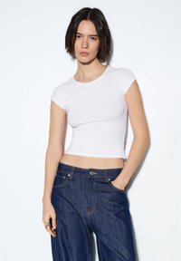 Bela obrezana majica s kratkimi rokavi, narejena iz gladke tkanine, v kombinaciji z visokim pasom temno modrih kavbojk z vidnim šivom in žepi.