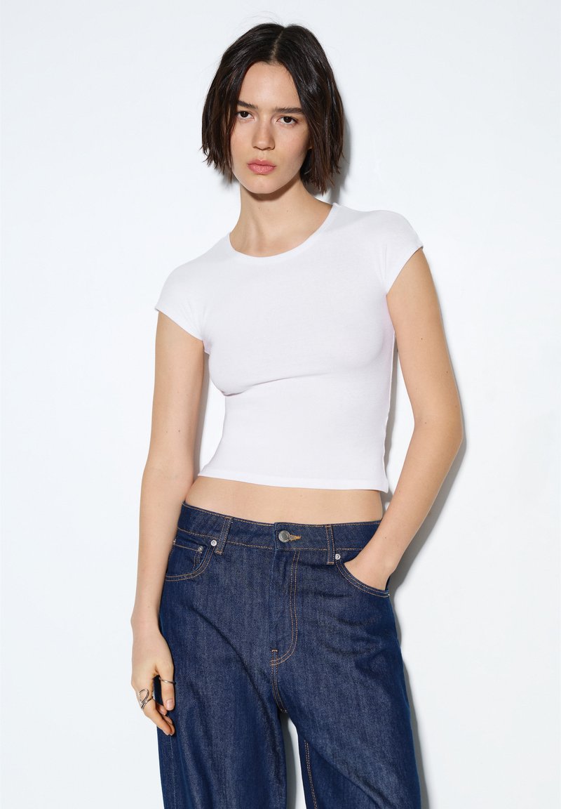 Bela obrezana majica s kratkimi rokavi, narejena iz gladke tkanine, v kombinaciji z visokim pasom temno modrih kavbojk z vidnim šivom in žepi.