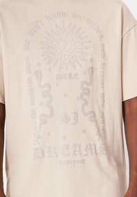 Camiseta de algodón beige con diseños gráficos texturizados, que incluyen un motivo de sol, serpientes y letras. Mangas cortas y corte relajado.