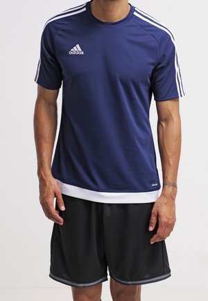 Homme portant un maillot de sport Adidas bleu marine avec des bandes blanches sur les manches et un short de sport noir, debout devant un fond uni.
