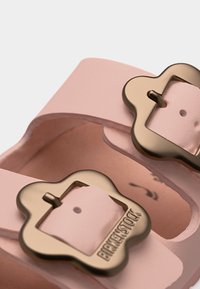 Κοντινό πλάνο ιμάντων σανδαλιών Birkenstock σε ανοιχτό ροζ χρώμα με μεταλλικά αγκράφια σε σχήμα λουλουδιού και ανάγλυφο όνομα της μάρκας σε μία από τις αγκράφες.