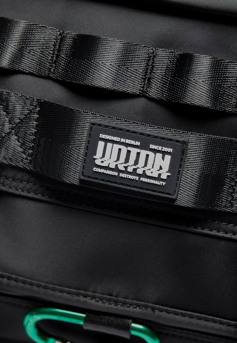 Interior de una bolsa de tela negra que presenta una correa texturizada con una etiqueta rectangular negra que dice "URBAN". Incluye un acento de mosquetón de metal verde.