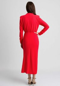 Czerwona dopasowana sukienka z długimi rękawami, wysoką szyją i wyraźnie zaznaczoną talią. Gładki materiał o długości midi, podkreślony detalem w postaci zamka.
