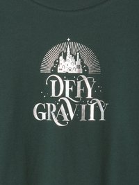 Sötétzöld pamut póló, ezüst színű grafikával, amelyen a "DEFY GRAVITY" felirat és egy kastély design található, felette sugárzó vonalakkal, sima textúrával.