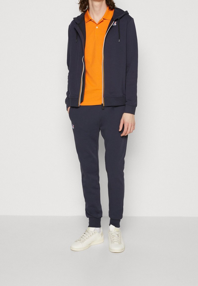 Personne portant un sweat à capuche zippé bleu marine et un pantalon de jogging par-dessus un polo orange avec des baskets blanches, debout devant un fond uni.