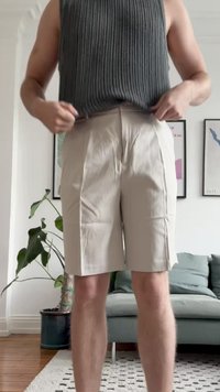 Beige Shorts mit Falten, aus leichtem Stoff gefertigt. Getragen mit einem dunkelgrauen ärmellosen Oberteil, das eine lockere Passform zeigt. Innenraum.