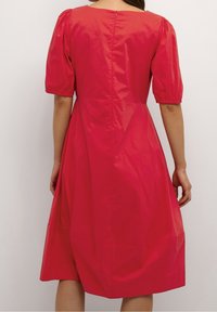 Nainen, joka on pukeutunut polvenpituiseen kirkkaan punaiseen mekkoon, jossa on kuppimaiset lyhyet hihat, kuvattuna takaa yksinkertaista vaaleaa taustaa vasten.