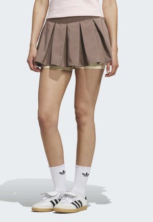 Osoba ubrana w brązową plisowaną spódnicę z beżowymi szortami pod spodem, białe skarpetki Adidas i białe tenisówki z czarnymi paskami.