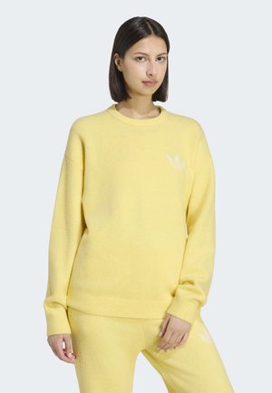 Kvinde med glat mørkt hår iført en gul langærmet sweatshirt og matchende bukser med subtile hvide logoer, stående mod en lys baggrund.