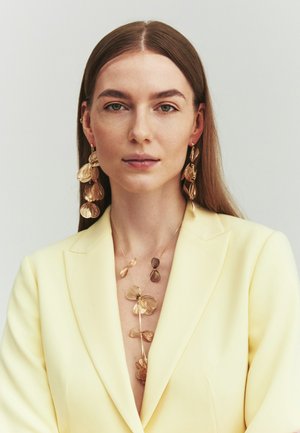 Woman with long brown hair wears large gold floral earrings and matching necklace paired with a pale yellow blazer, looking directly forward.