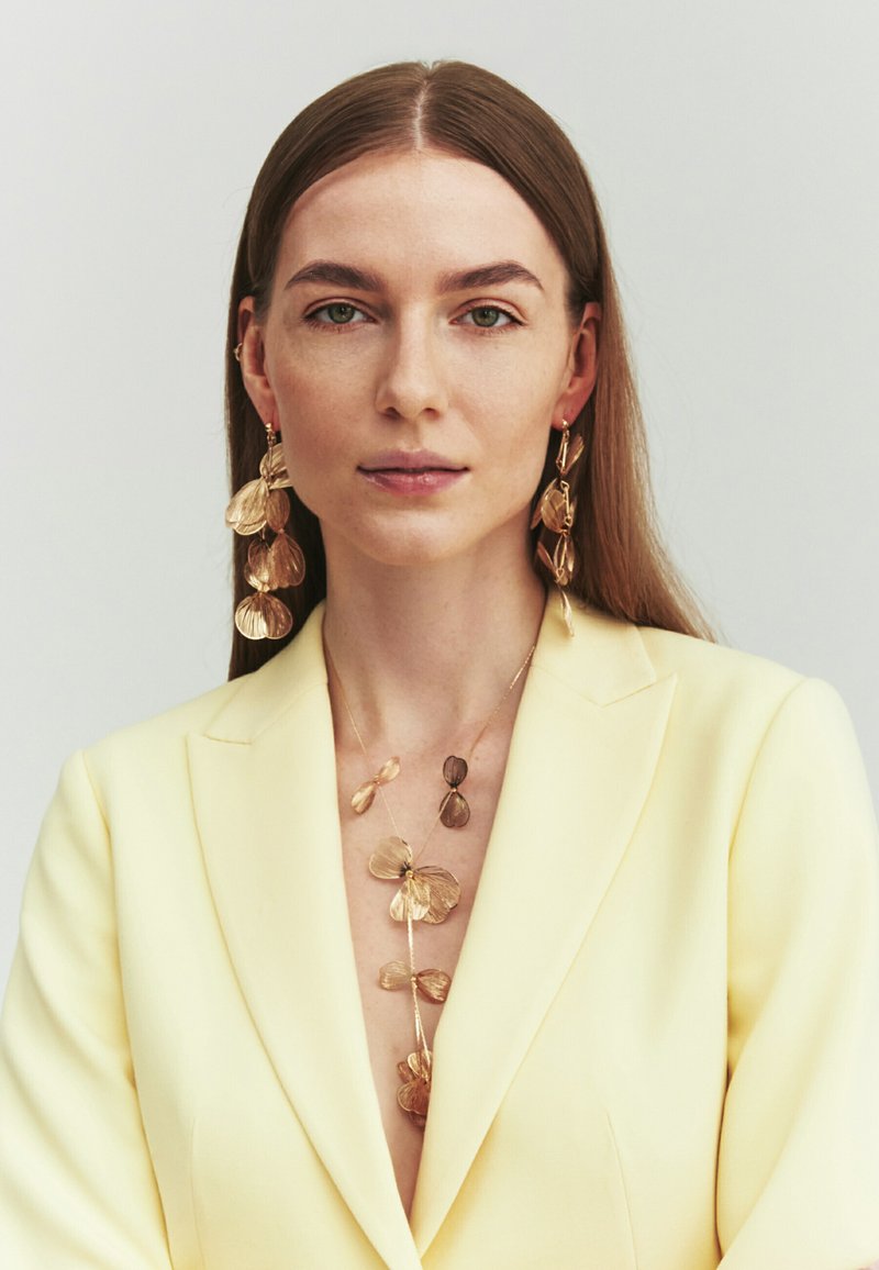Femme aux longs cheveux bruns porte de grandes boucles d’oreilles florales en or assorties d’un collier, associées à un blazer jaune pâle, regardant droit devant.