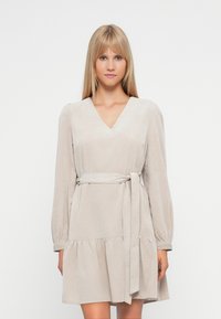 Svetlo bež obleka z V-izrezom, dolgimi rokavi in volanastim robom. Ima tkaninski pas in teksturiran material za dodaten detajl.