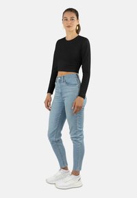 Schwarzes, kurzes Langarmoberteil, hellblaue High-Waist-Jeans und weiße Sneaker. Das Modell trägt gelbe Ohrringe. Glattes, schmiegsames Material und lässiger Stil.