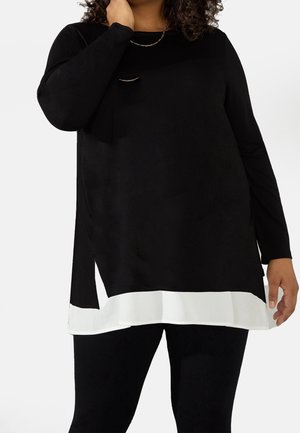 Frau trägt eine schwarze Langarm-Tunika mit einem weißen Saumdetail und schwarzen Leggings, posiert mit einer Hand in der Nähe ihres Halses.