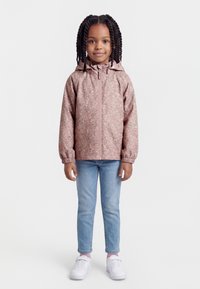 Fată tânără cu părul împletit, purtând o geacă roz cu imprimeu floral și glugă, blugi deschiși la culoare și adidași albi, stând cu fața spre înainte.