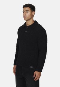 Maglione nero a trama con collo, tre bottoni e orlo dritto. Indossato con pantaloni neri, che presentano un'etichetta con logo all'orlo.