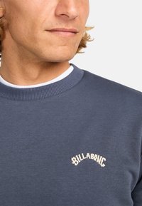 Sudadera azul marino con cuello acanalado y tejido texturizado. Presenta un logo bordado de color crema en el pecho.