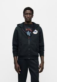 Μαύρο φούτερ με φερμουάρ μπροστά, με μπροστινή τσέπη και έμβλημα στο αριστερό στήθος. Φορώντας πάνω από ένα γραφικό t-shirt, σε συνδυασμό με μαύρο παντελόνι.