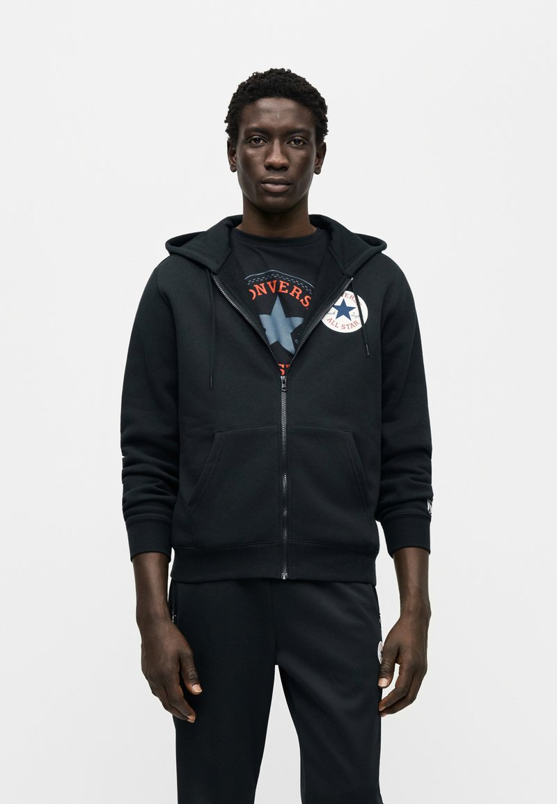 Sort hættejakke med lynlås foran og en lomme foran samt et emblem på venstre bryst. Båret over en grafisk t-shirt, kombineret med sorte bukser.