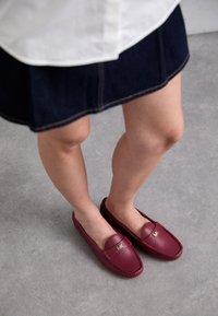 Maroon leather loafers with a flat design, featuring a gold emblem on the front, paired with a dark denim skirt and light-colored background.