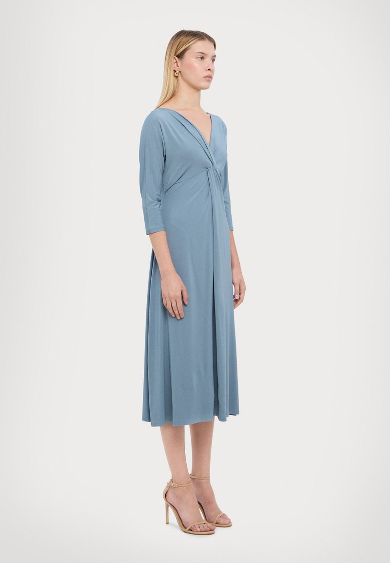 Blauwe jurk tot de knie met een diepe V-hals, draai-detail bij de taille en driekwart mouwen. Draagt met beige strap-sandalen met hoge hakken.