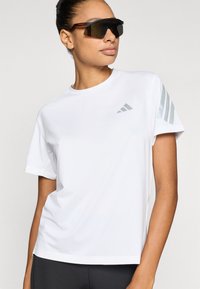 Fehér sportpóló kerek nyakkivágással és rövid ujjal. A mellkason szürke logó, a bal ujjon pedig három szürke csík található.