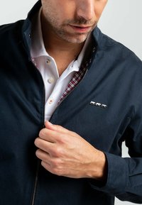 Veste marine avec fermeture éclair sur le devant, doublure à carreaux contrastée et petite broderie d'animal sur la poitrine. Texture de tissu lisse.