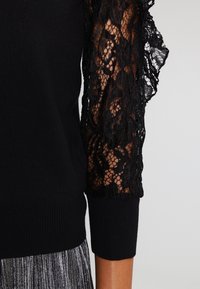 Černý pletený top s propracovanými květinovými krajkovými rukávy, které odhalují pleť, doplněný skladanou metalickou stříbrnou sukní.