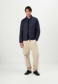 Tamsiai mėlynas pūkinis švarkas su užtrauktuku, turi ribotą apykaklę, su siuvinėta tekstūra, dėvimas ant tamsiai mėlynos marškinių, derinamas su smėlio spalvos kelnėmis ir juodais batais.