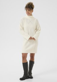 Abito a maglia color crema con motivo a trecce, colletto alto, maniche lunghe e vestibilità slim. Abbinato a stivali in pelle nera al ginocchio.