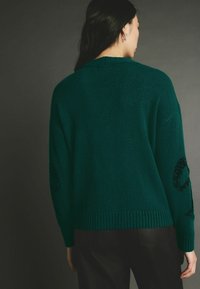 Pull en teal avec un ourlet côtelé et des manches longues, doté d'embellissements circulaires texturés sur les coudes. Matière douce en tricot.