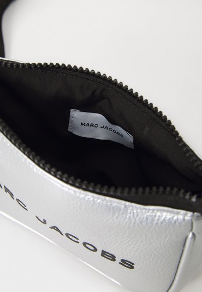 Sølv glidelåsveske med en teksturert overflate, med svart innside og en hvit etikett som viser "MARC JACOBS."