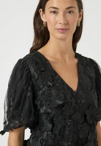 Zwarte jurk met bloemenborduursel, V-hals en opgeblazen, doorschijnende mouwen. De stof heeft een zachte textuur en ingewikkelde bloemmotieven.