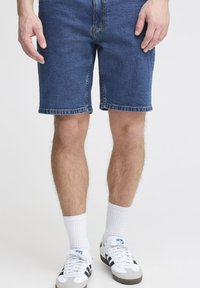Homme portant un short en jean bleu, des chaussettes blanches hautes et des baskets blanches à rayures noires, debout devant un fond uni.