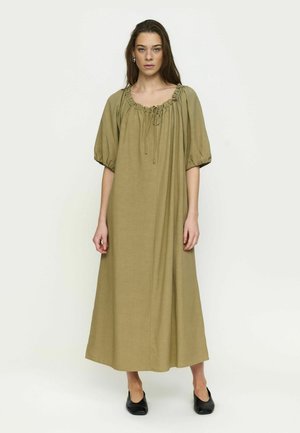 Vrouw draagt een losse, olijfgroene jurk tot op de enkels met pofmouwen en een geplooide halslijn, staand met zwarte platte schoenen tegen een witte achtergrond.