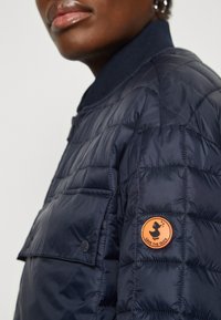 Marine quiltet pufferjakke med høy krage, har en frontlomme og en oransje sirkulær logo-lapp på venstre erme.