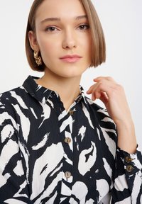 Schwarz-weiß gemusterte Bluse mit einem Kragen zum Knöpfen, strukturiertem Stoff und braunen Knöpfen. Das Model hat kurze Haare und trägt goldene Hoop-Ohrringe.