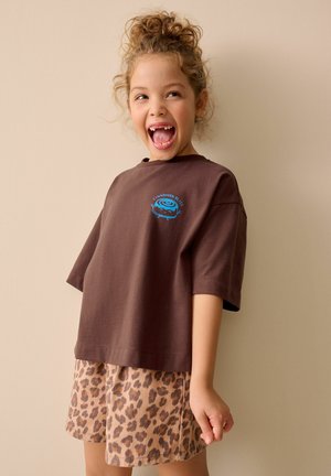 Jong meisje met krullend haar dat breed glimlacht, gekleed in een bruin T-shirt met een blauwe kaneelbroodjesafbeelding en korte broek met luipaardprint.