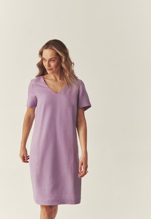 Kobieta w krótkim, sięgającym do kolan liliowym sukience z krótkim rękawem i dekoltem w kształcie litery V, stojąca na jednolitym jasnym tle.
