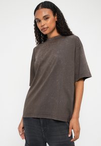 Kvinde med krøllet hår iført en løs mørkegrå kortærmet skjorte og mørk jeans, der står foran en hvid baggrund.