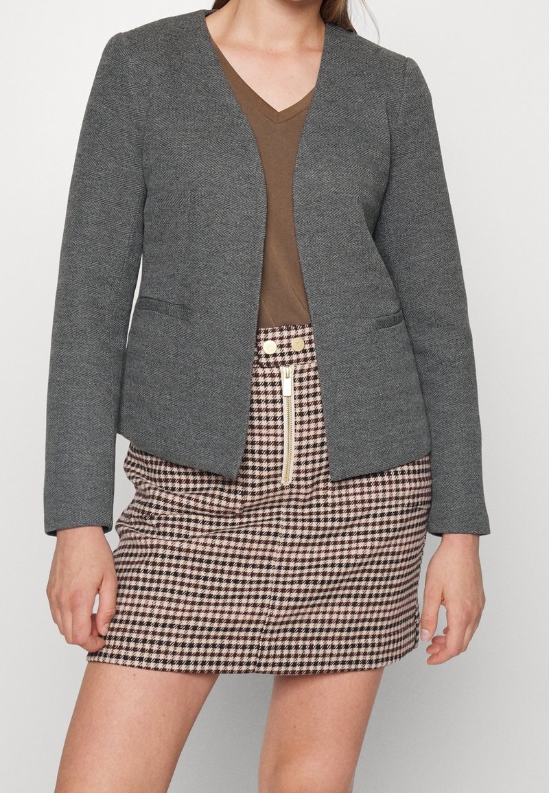 Blazer gris à chevrons avec un col en V, présentant deux poches avant, assorti à une chemise marron et une jupe à carreaux dotée d'une fermeture éclair à l'avant.
