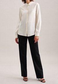 Woman wearing a cream buttoned long-sleeve blouse, black straight-leg trousers, and black open-toe heels standing on a neutral background.