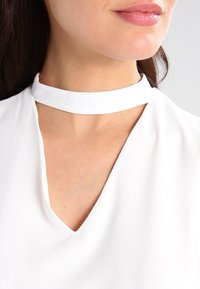 Blusa blanca con escote en V y un amplio collar tipo choker. El material es liso con una textura sutil. Sin patrones ni adornos visibles.