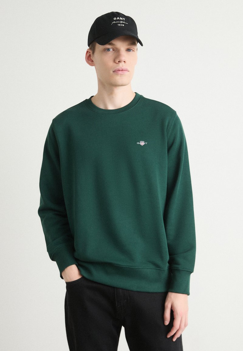 Groene sweatshirt met lange mouwen en rib gebreide boorden, met een klein geborduurd logo op de borst. Gecombineerd met een zwarte pet.