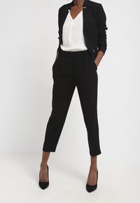 Blazer noir cintré porté sur un chemisier blanc à col en V, accompagné d'un pantalon noir sur mesure et de chaussures à talons noires. Textures lisses et design moderne.